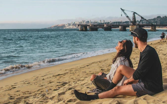 Couple sitting on the beach near Muelle Vergara, Viña del Mar, with historic crane in view.