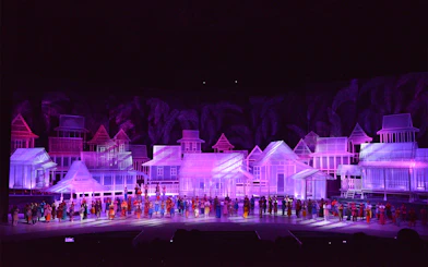 Traditional village scene with performers in colorful attire on stage, Kampung Scen.