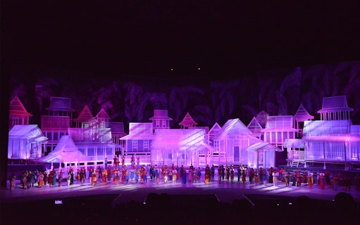 Traditional village scene with performers in colorful attire on stage, Kampung Scen.