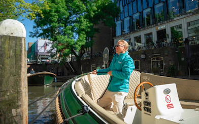 Boat captain steering a canal cruise in Utrecht, Netherlands, with city buildings in the background.