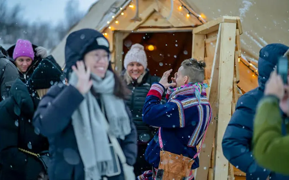 Sami guide in traditional attire outside a wooden tent in snowy Tromsø.