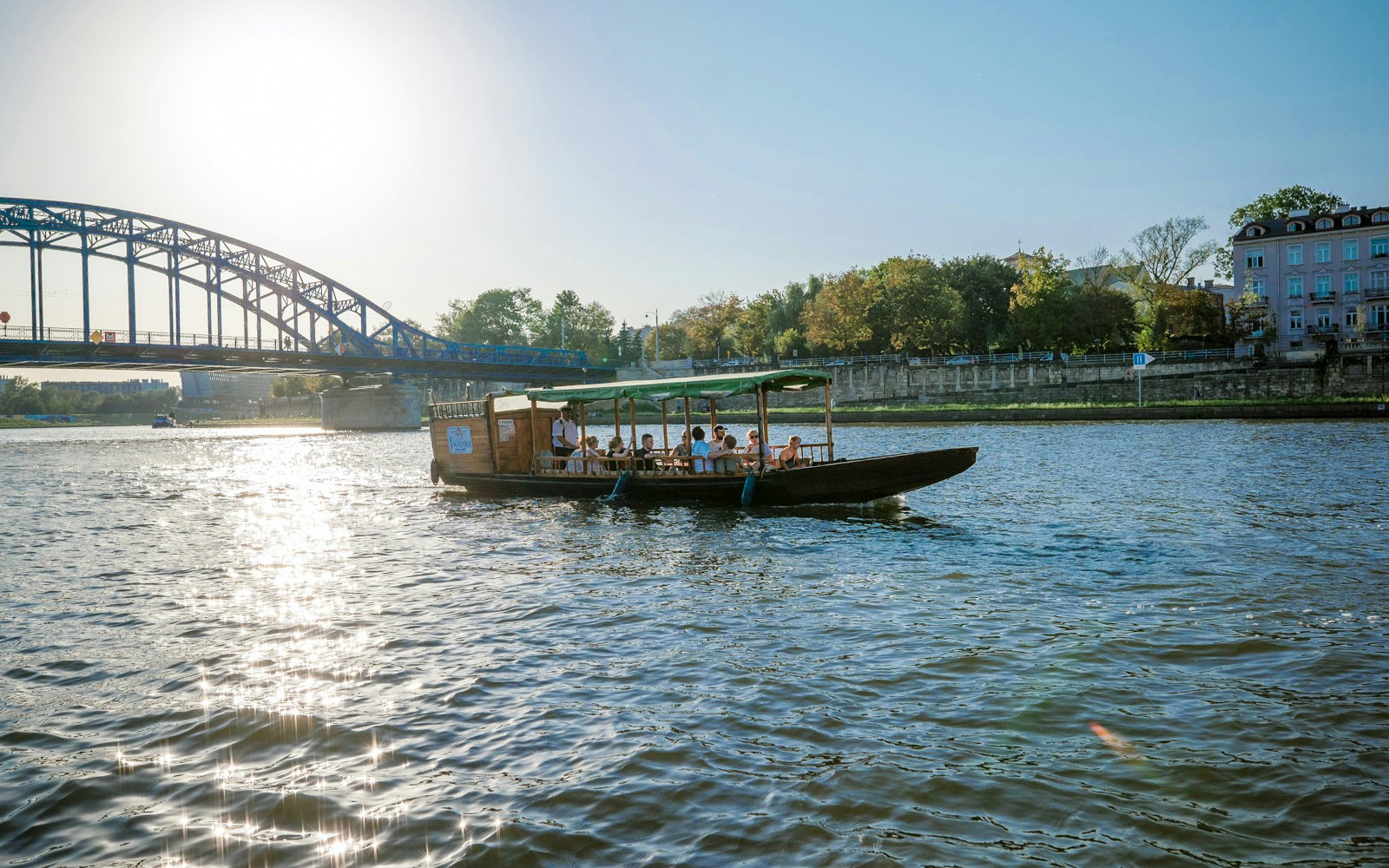 Boat cruising on Vistula River with passengers, bridge and cityscape in background.