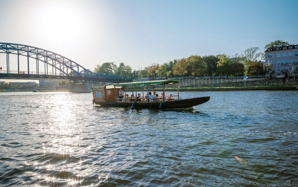 Boat cruising on Vistula River with passengers, bridge and cityscape in background.