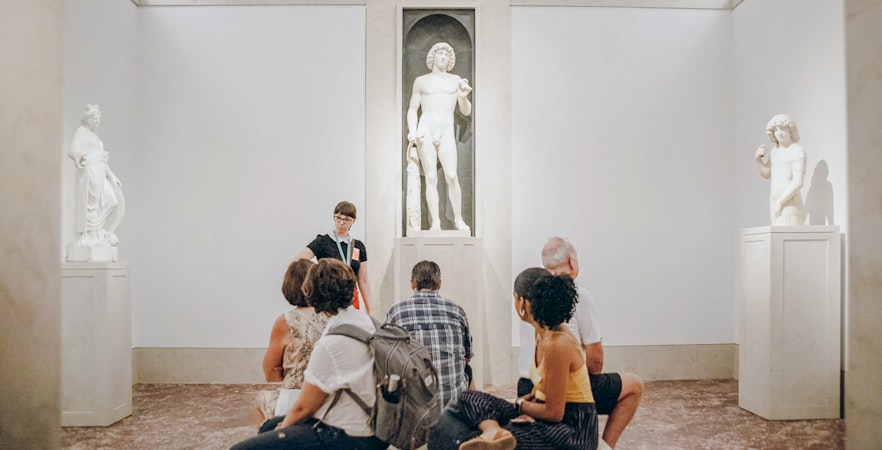 Tour group seated on bench listening to guide near marble nude statue at Metropolitan Museum of Art, New York.