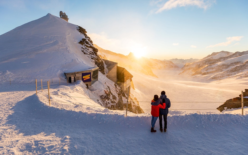 Couple admiring sunrise at Jungfraujoch Top of Europe, snowy mountain landscape.
