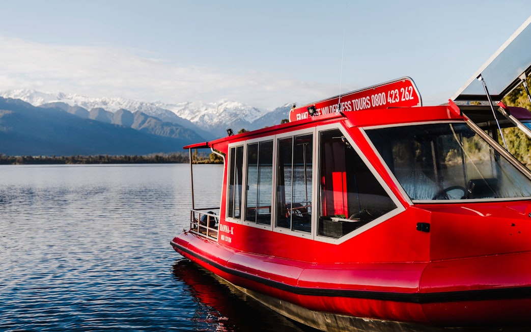 Red tour boat on Lake Mapourika with snow-capped mountains in the background.