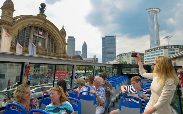 Tourists on a hop-on hop-off bus near Frankfurt Hauptbahnhof with city skyline in view.