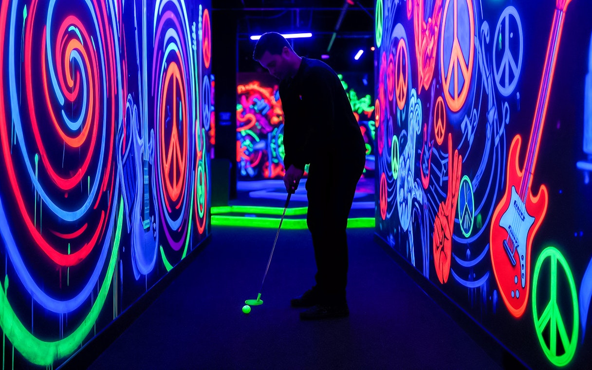 Man playing minigolf in a neon-lit Rock of Ages blacklight course with vibrant wall art.