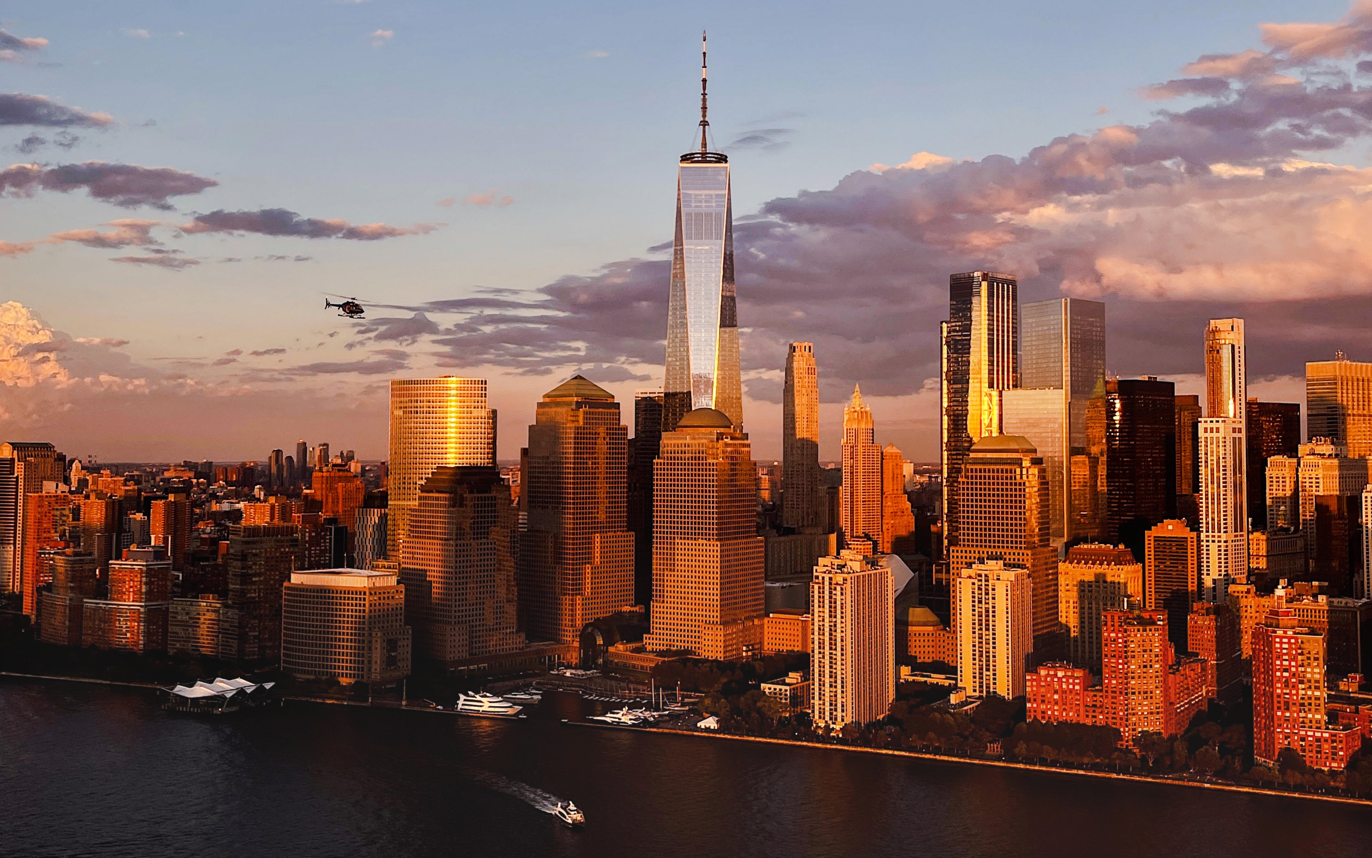 Helicopter flying over New York City skyline with One World Trade Center at sunset.