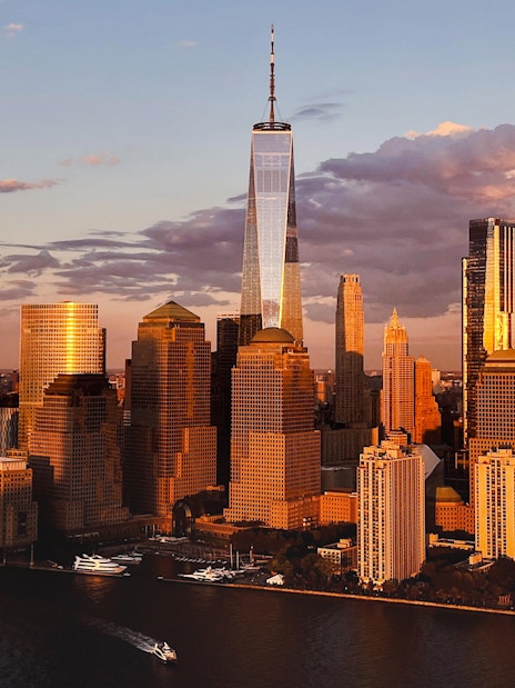 Helicopter flying over New York City skyline with One World Trade Center at sunset.