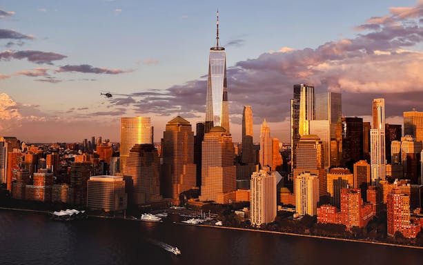 Helicopter flying over New York City skyline with One World Trade Center at sunset.