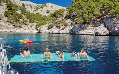 People relaxing on a floating mat and kayaking near rocky cliffs during a catamaran trip in Alcudia.
