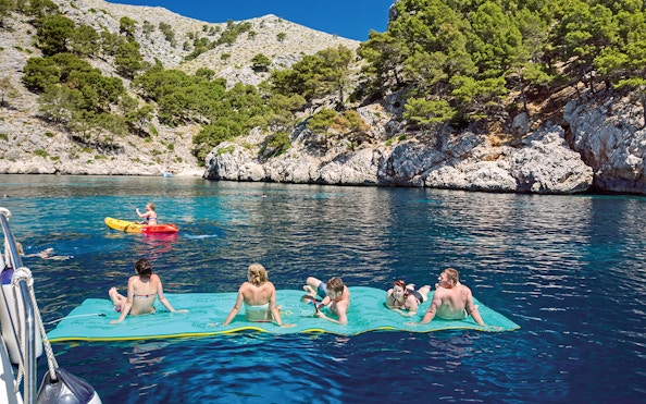 People relaxing on a floating mat and kayaking near rocky cliffs during a catamaran trip in Alcudia.