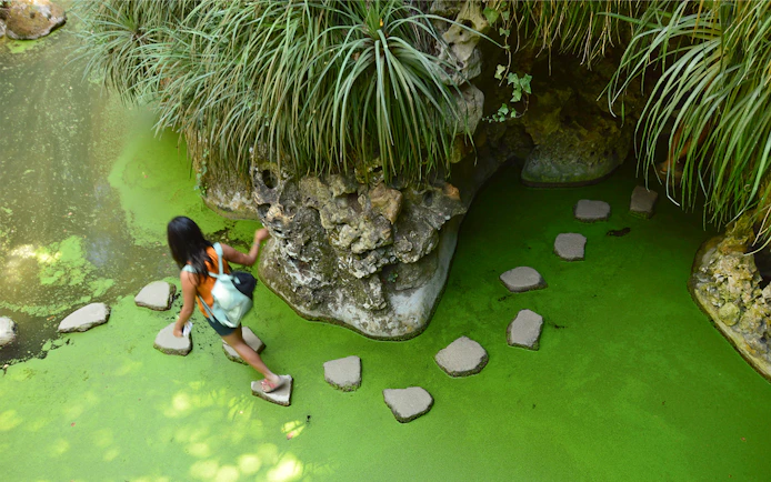 Girl walking on stepping stones over a green pond in a lush garden.
