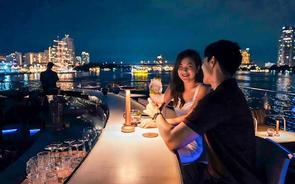 Couple enjoying evening drinks on Saffron Luxury Cruise by Banyan Tree with city skyline.