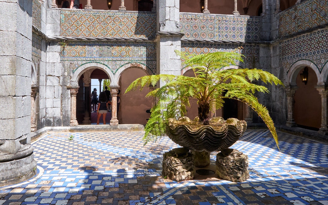 Cloisters with ornate arches and a central fern in a shell-shaped planter.