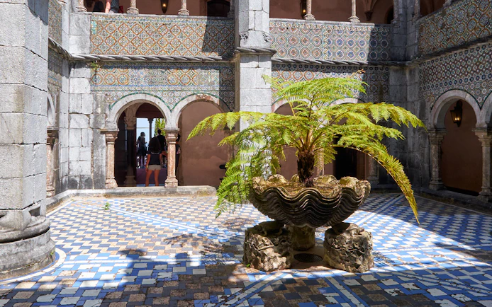 Cloisters with ornate arches and a central fern in a shell-shaped planter.
