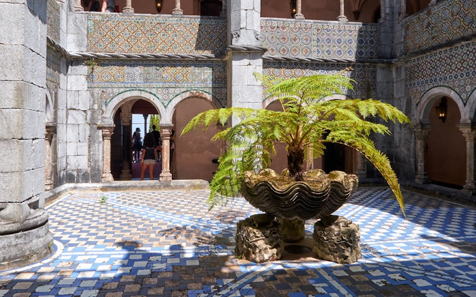 Cloisters with ornate arches and a central fern in a shell-shaped planter.