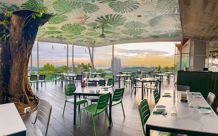 Outdoor dining area with view of Singapore skyline at sunset, featuring lush greenery and modern decor.