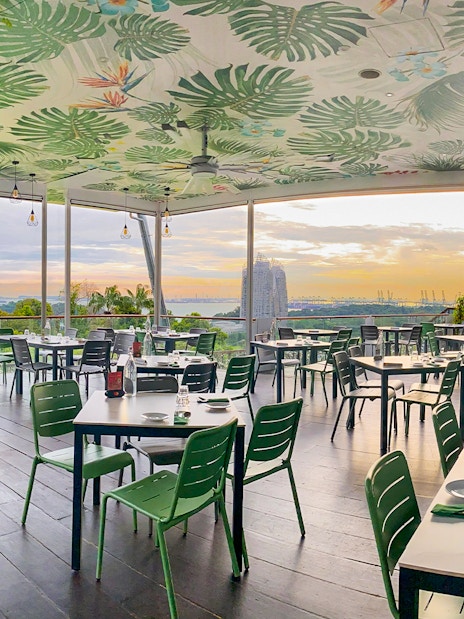 Outdoor dining area with view of Singapore skyline at sunset, featuring lush greenery and modern decor.