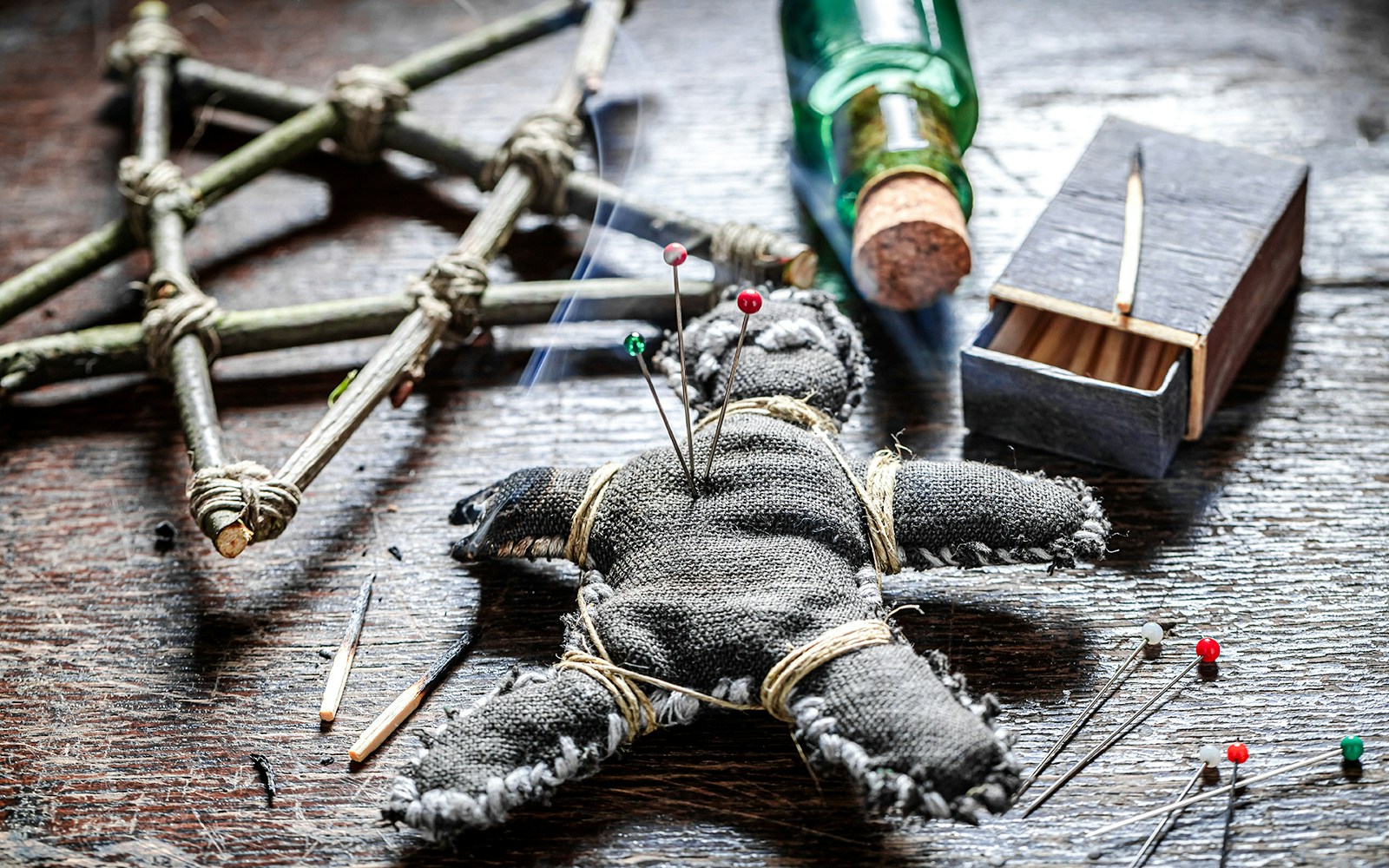 Voodoo doll with pins on a wooden table, surrounded by a pentagram and bottle.