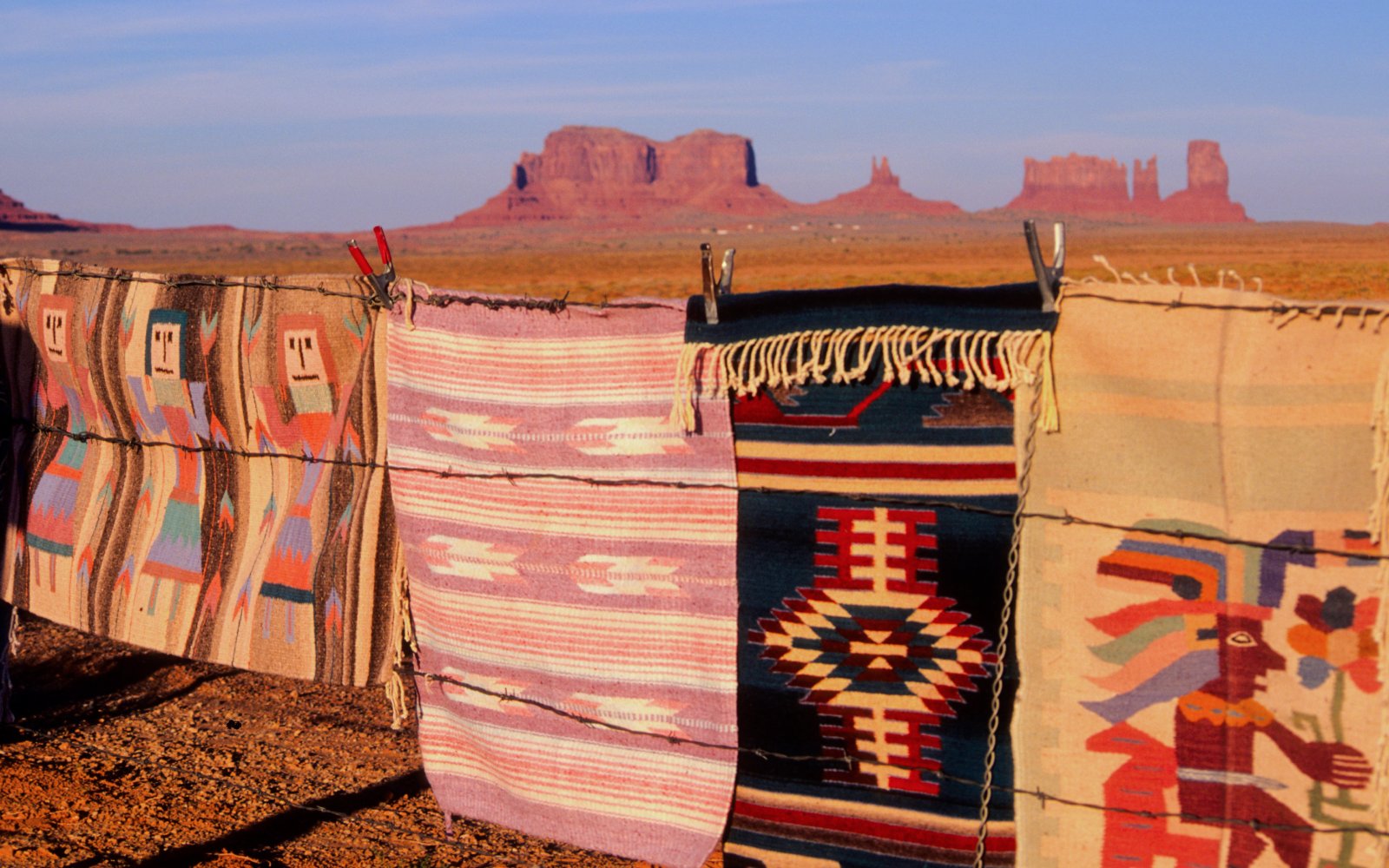 Traditional Navajo rugs on barbed wire with Monument Valley buttes in the background.