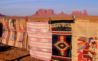 Traditional Navajo rugs on barbed wire with Monument Valley buttes in the background.