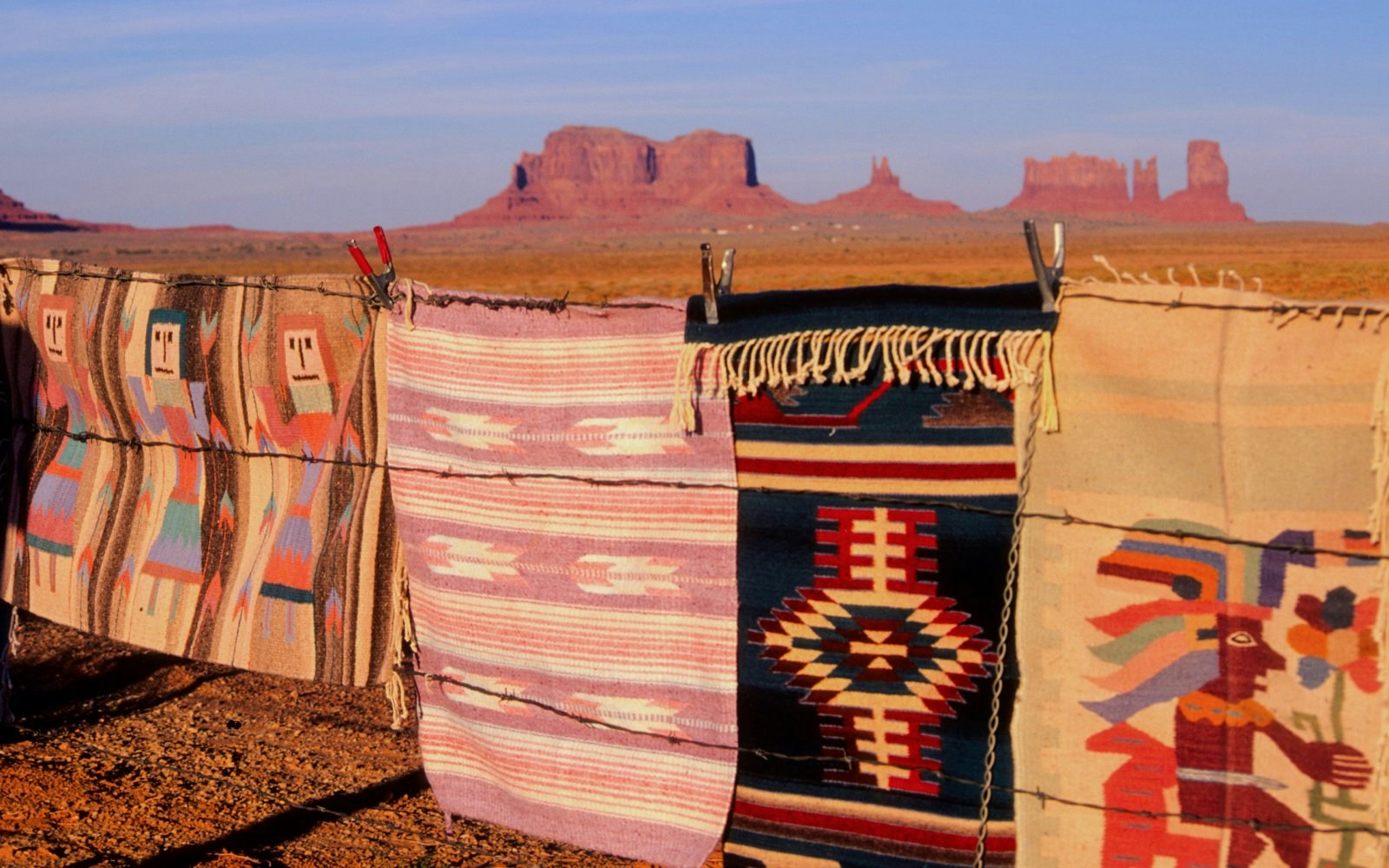 Traditional Navajo rugs on barbed wire with Monument Valley buttes in the background.