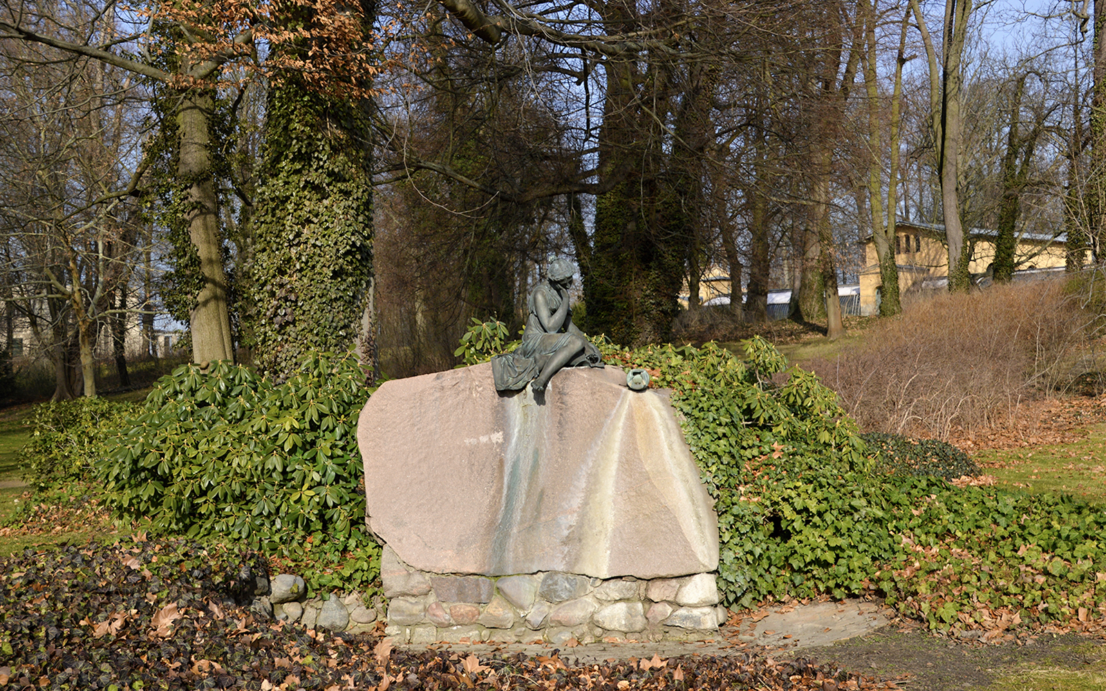 Statue of a seated figure on a rock in Glienicke Park, Germany, surrounded by trees and foliage.
