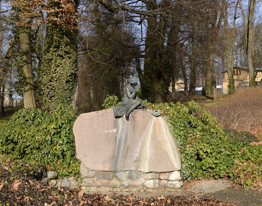 Statue of a seated figure on a rock in Glienicke Park, Germany, surrounded by trees and foliage.