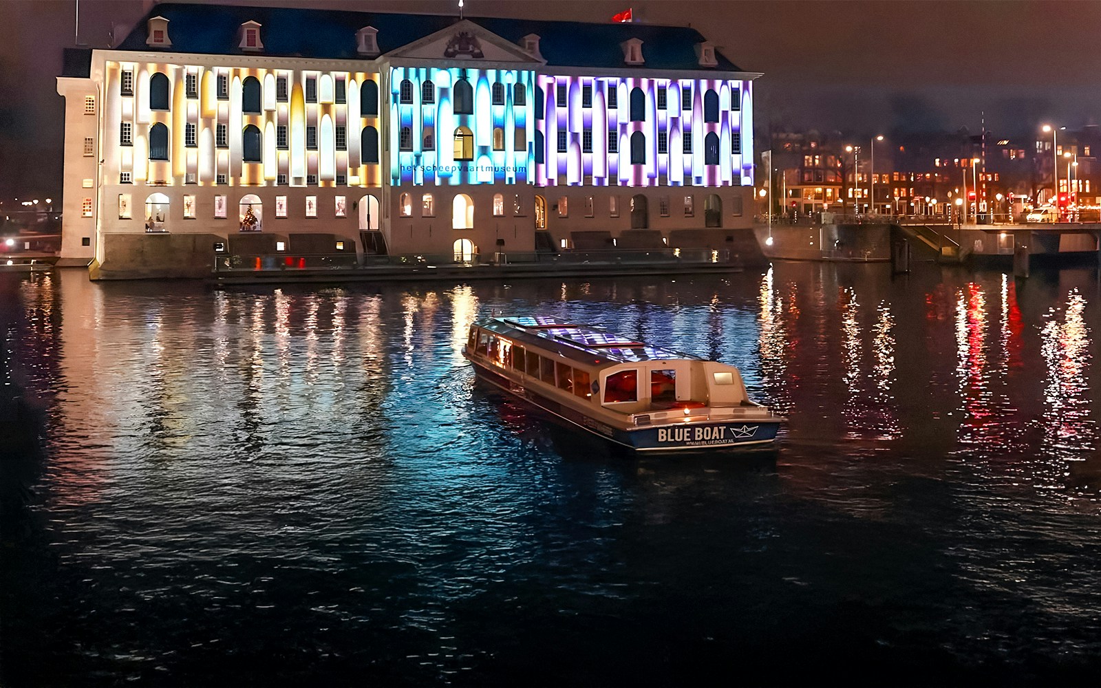 Evening cruise boat on Amsterdam canal with illuminated building during Light Festival.
