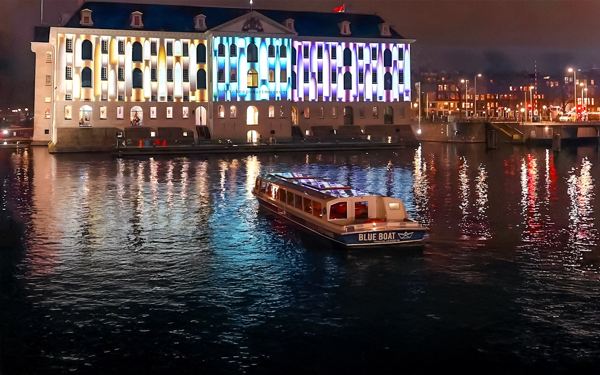 Evening cruise boat on Amsterdam canal with illuminated building during Light Festival.