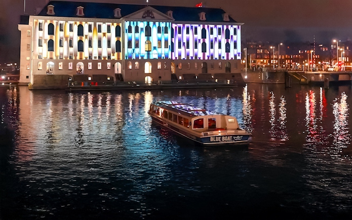 Evening cruise boat on Amsterdam canal with illuminated building during Light Festival.