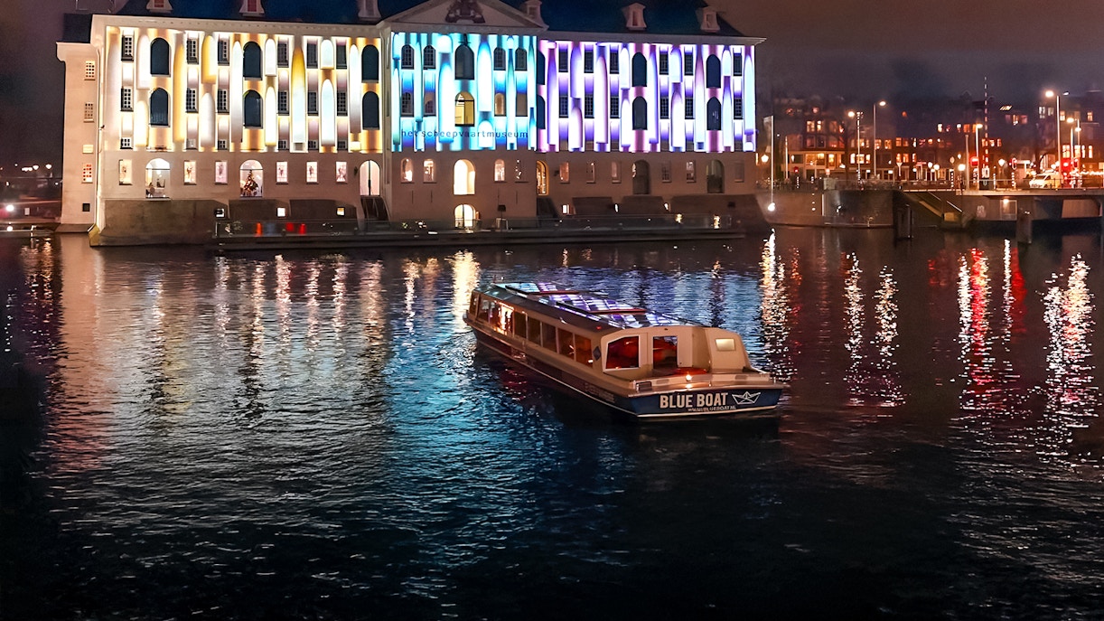 Evening cruise boat on Amsterdam canal with illuminated building during Light Festival.