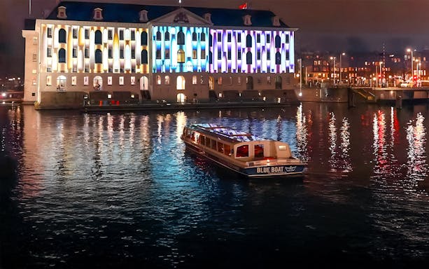 Evening cruise boat on Amsterdam canal with illuminated building during Light Festival.