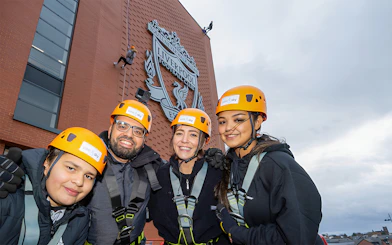 Group preparing for abseil at Liverpool FC's Anfield Stadium with club emblem in background.