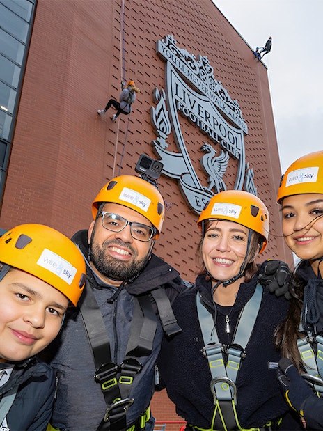 Group preparing for abseil at Liverpool FC's Anfield Stadium with club emblem in background.