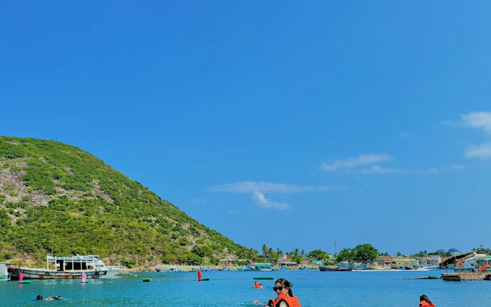 Tourists swimming near boats and lush hills on Robinson Beach, Nha Trang.