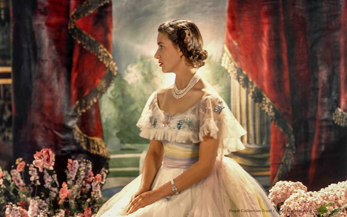 A woman in an elegant gown with pearls, seated in a regal setting with red drapes and flowers.