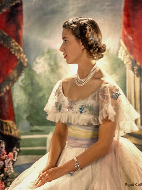 A woman in an elegant gown with pearls, seated in a regal setting with red drapes and flowers.