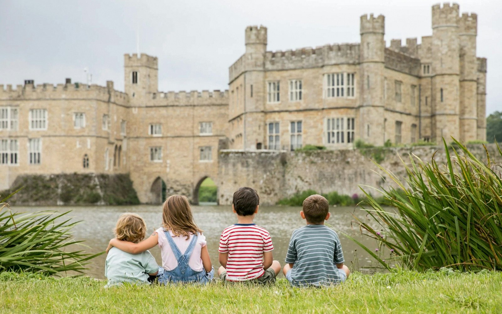 Children sitting on grass facing Leeds Castle across the moat.