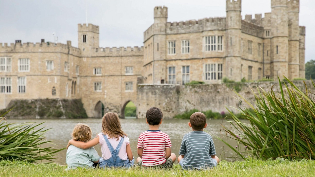 Children sitting on grass facing Leeds Castle across the moat.