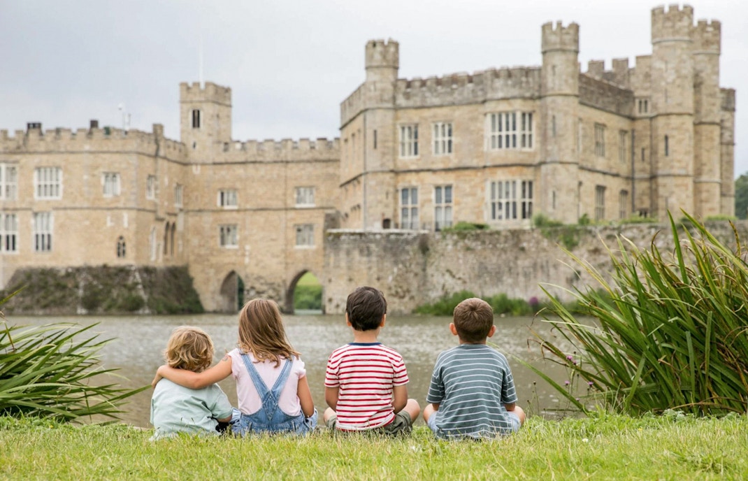 Children sitting on grass facing Leeds Castle across the moat.