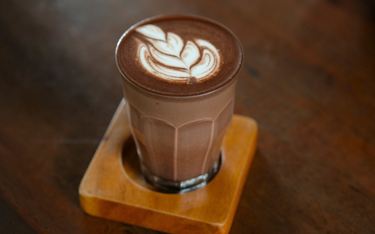 Glass of hot chocolate with latte art on a wooden coaster.