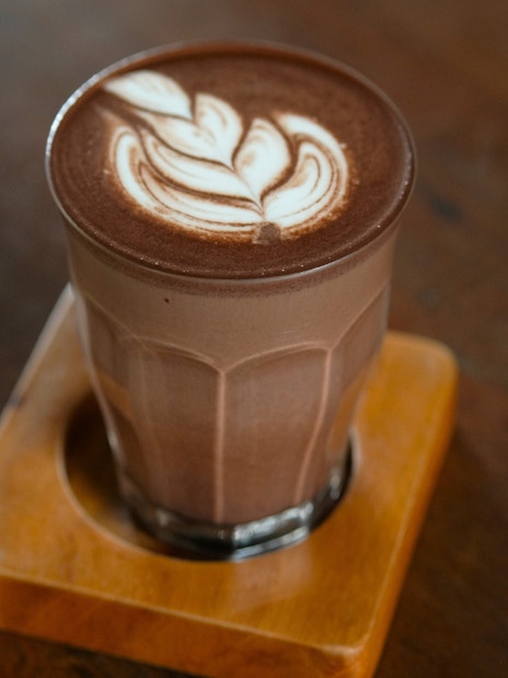 Glass of hot chocolate with latte art on a wooden coaster.