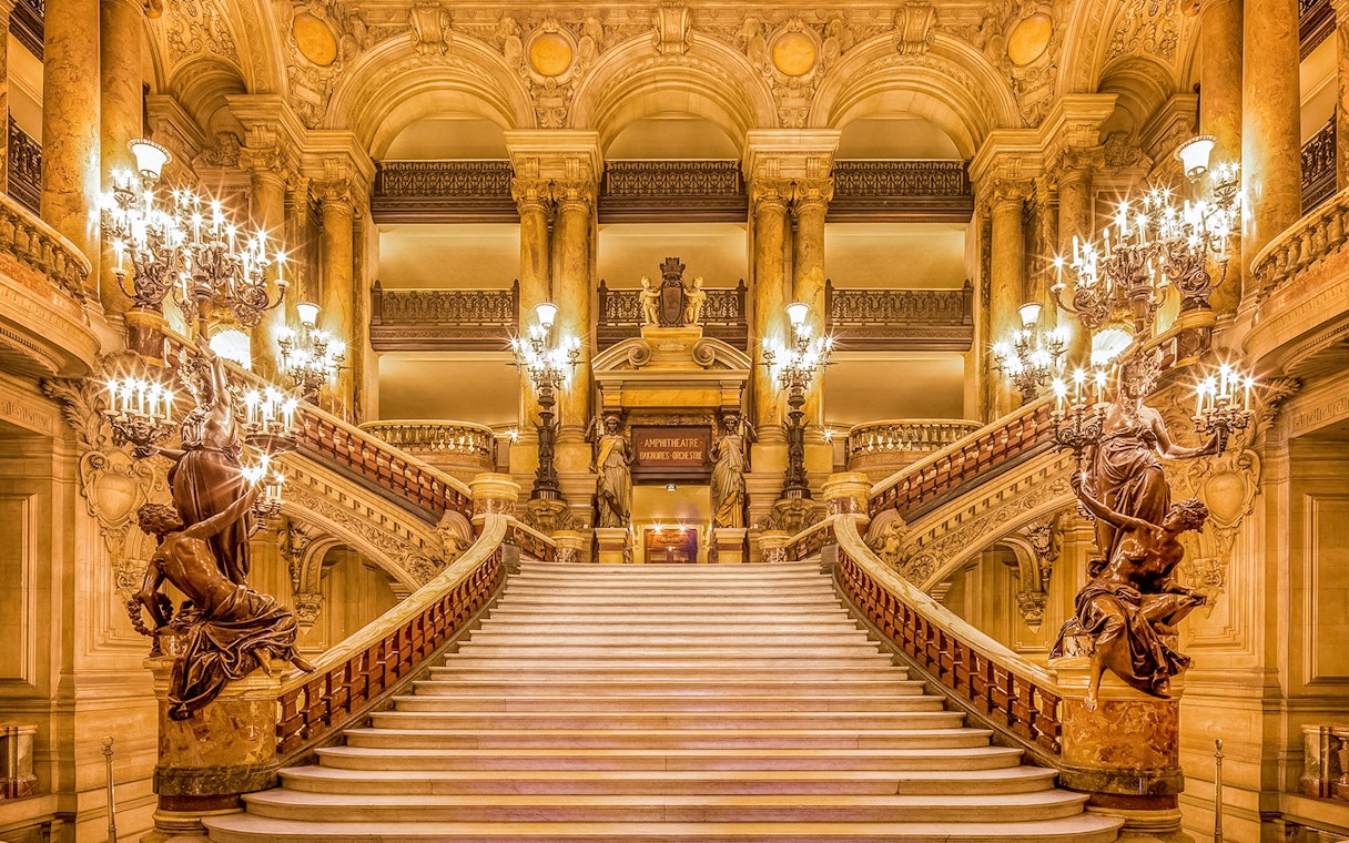 Grand staircase inside Opéra Garnier, Paris, with ornate chandeliers and sculptures.