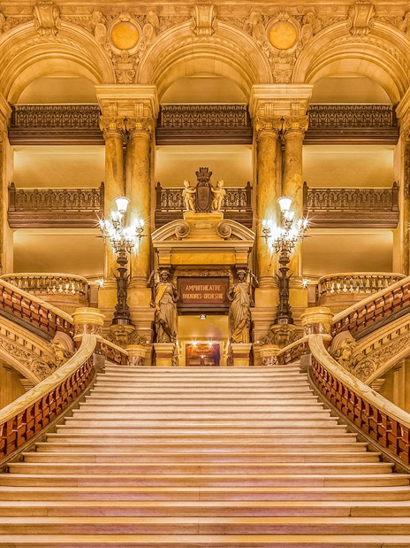 Grand staircase inside Opéra Garnier, Paris, with ornate chandeliers and sculptures.