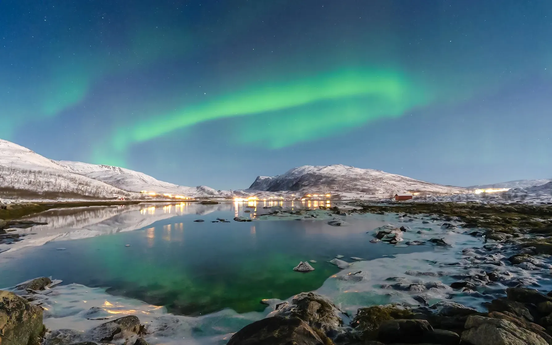 Northern lights over snowy mountains and a reflective lake in Tromso, Norway.
