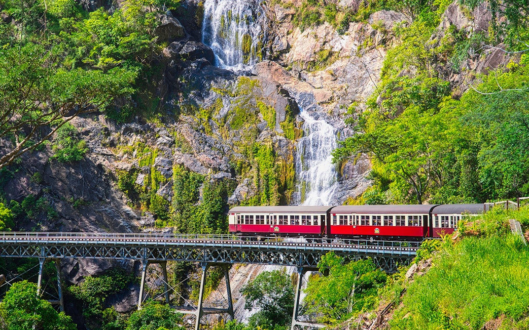 Kuranda Scenic Railway train crossing bridge with waterfall backdrop in Queensland, Australia.