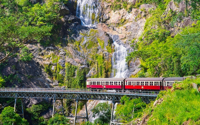 Kuranda Scenic Railway train crossing bridge with waterfall backdrop in Queensland, Australia.